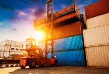 Άνοιγμα κοντέινερ σε τελωνειακό χώρο με φορτωτή και στοιβαγμένα εμπορευματοκιβώτια