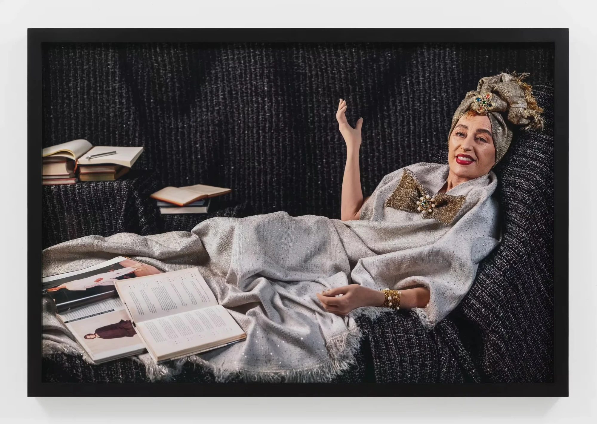 Η Cindy Sherman ξαπλωμένη με γκρι ένδυμα μπροστά σε μαύρο φόντο, με ανοιχτά βιβλία και περιοδικά