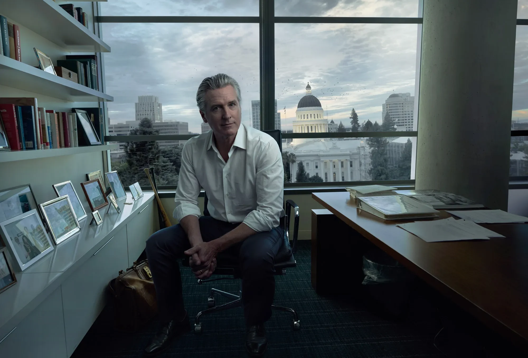 Ο Gavin Newsom καθισμένος μπροστά σε παράθυρο γραφείου με το Καπιτώλιο της Καλιφόρνιας στο βάθος.