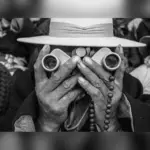 Ασπρόμαυρη φωτογραφία προσώπου με καπέλο που κρατά κιάλια μπροστά στα μάτια του, ανάμεσα σε άλλους ανθρώπους.