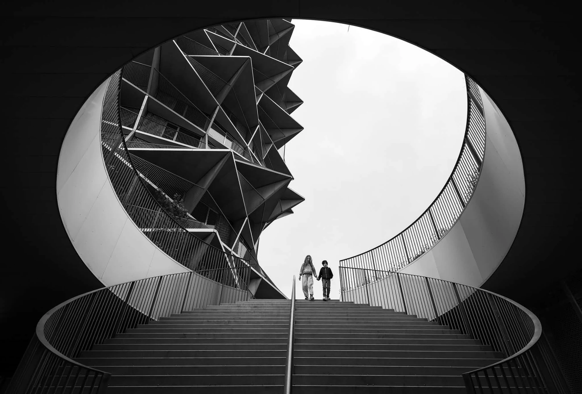 Black-and-white architectural photograph of a curved staircase, geometric buildings and two people