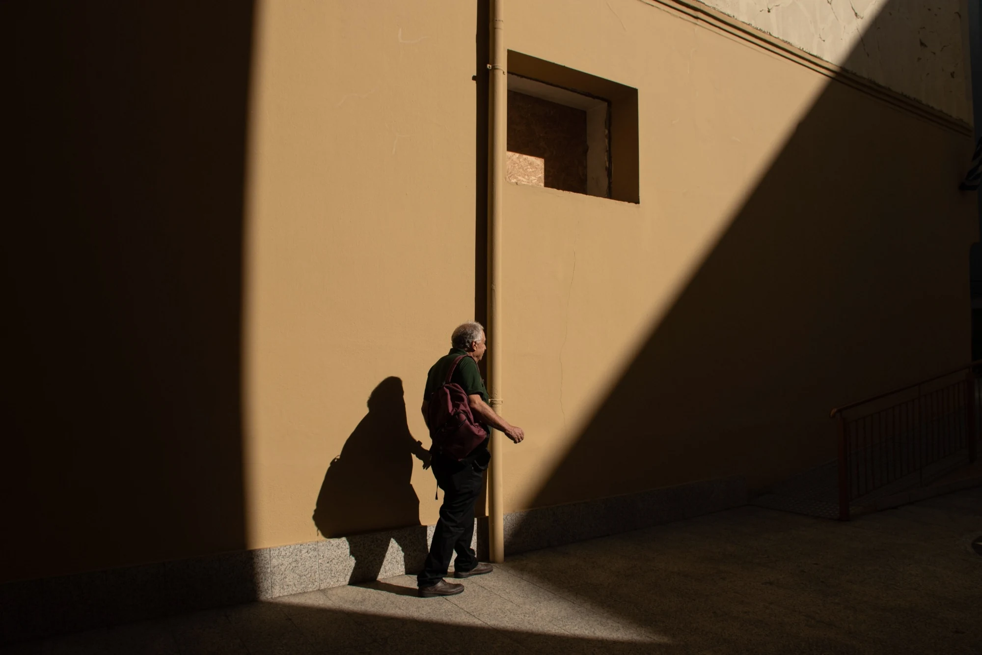Άνδρας περπατά μπροστά σε τοίχο με έντονο φως και γεωμετρικές σκιές.